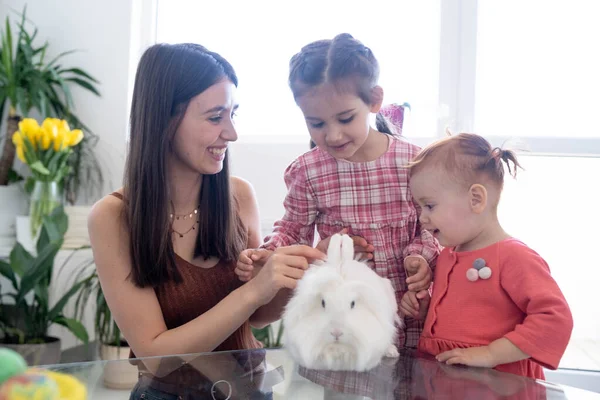 Cheerful smiley mother and her daughters playing with Easter white rabbit and laughing. Happy family preparing for Easter. Easter family concept. White window background. Yellow flowers.