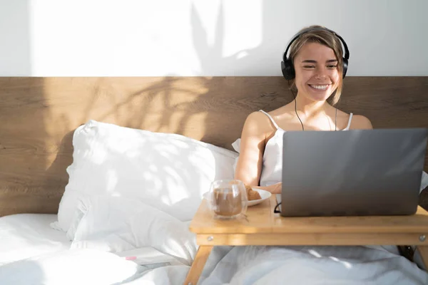 Smiling young woman in earphones using laptop, listening music and eating croissants with coffee for breakfast. Happy female communicating by video conference call sit on bed at home.
