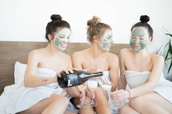 Beautiful smiling multicultural girls in treatment masks celebrating with champagne at home during pajama party.  Three pretty female friends drinking champagne and laughing. Spa self care concept.