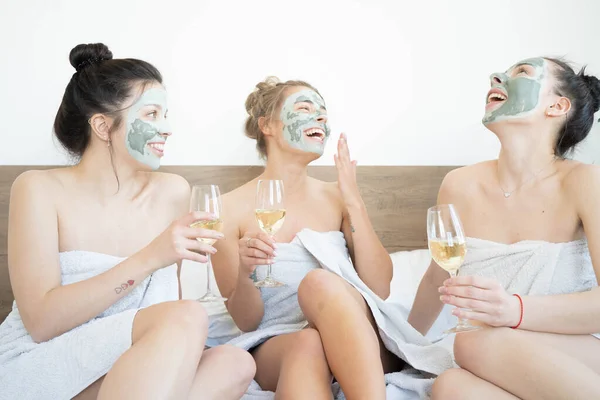 Beautiful smiling multicultural girls in treatment masks celebrating with champagne at home during pajama party.  Three pretty female friends drinking champagne and laughing. Spa self care concept.