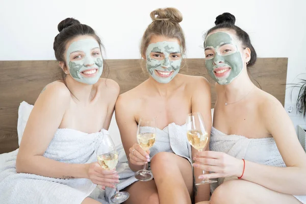 Beautiful smiling multicultural girls in treatment masks celebrating with champagne at home during pajama party.  Three pretty female friends drinking champagne and laughing. Spa self care concept.