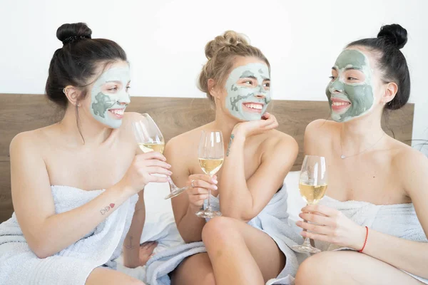 Beautiful smiling multicultural girls in treatment masks celebrating with champagne at home during pajama party.  Three pretty female friends drinking champagne and laughing. Spa self care concept.