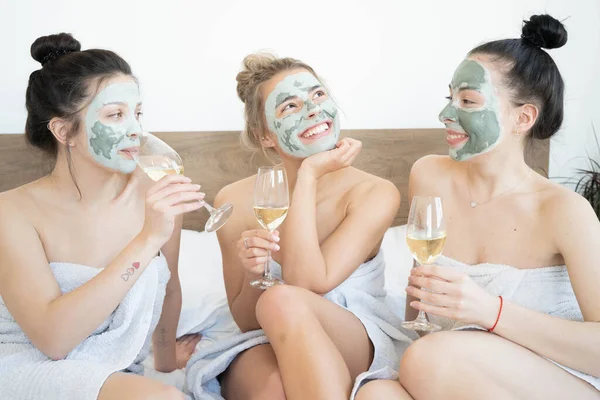 Beautiful smiling multicultural girls in treatment masks celebrating with champagne at home during pajama party.  Three pretty female friends drinking champagne and laughing. Spa self care concept.