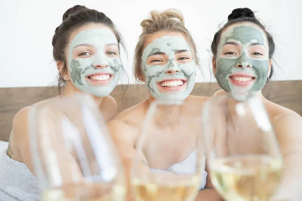 Beautiful smiling multicultural girls in treatment masks celebrating with champagne at home during pajama party.  Three pretty female friends drinking champagne and laughing. Spa self care concept.