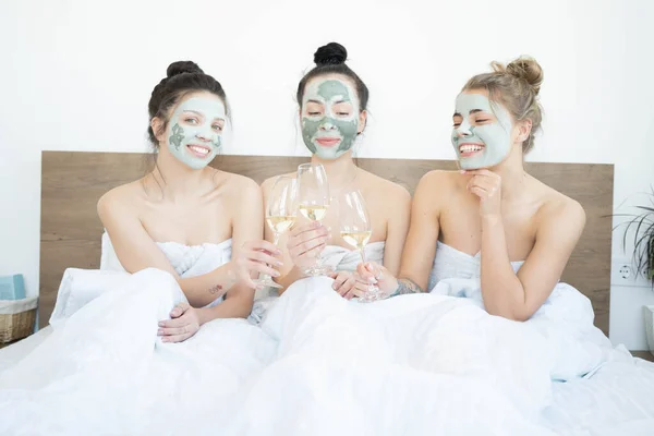 Beautiful smiling multicultural girls in treatment masks celebrating with champagne at home during pajama party.  Three pretty female friends drinking champagne and laughing. Spa self care concept.