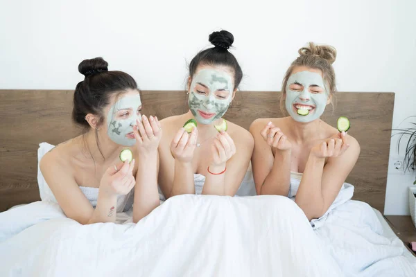 Three funny happy beautiful girlfriends making face clay masks, laughing and eating cucumbers. Stay at home concept. Skin care and treatment, spa, natural beauty and cosmetology concept.
