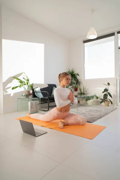 Young beautiful woman practicing yoga and gymnastic. Attractive girl practicing stretching and yoga at home. Woman doing body exercise near computer in morning. Healthy and sport lifestyle. Home workout.