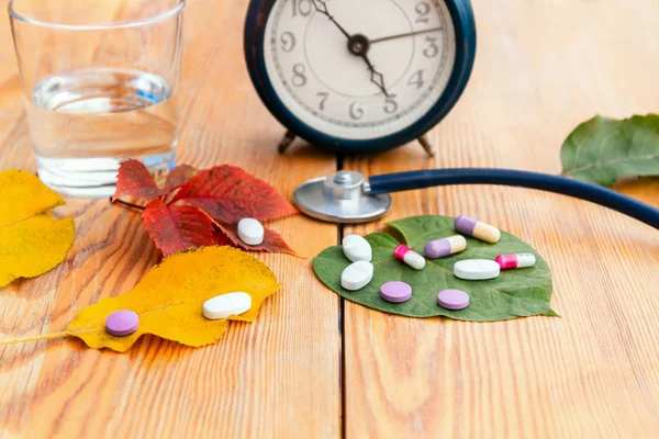 Autumn colds. Time to take pills. Alarm clock, stethoscope, colorful pills, autumn leaves of different colors on a wooden table.