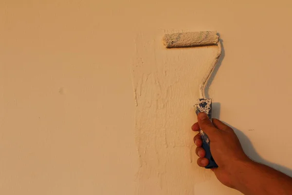 A male right hand is painting the white wall with paint roller and white dye.