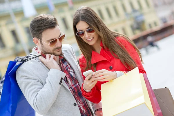 Jonge gelukkige paar met slimme telefoon en shopping tassen in de stad — Stockfoto