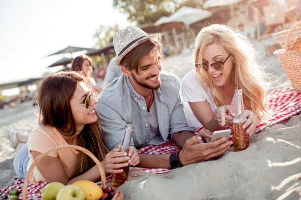 One man and two women lying on the beach and looking into phone and smiling.