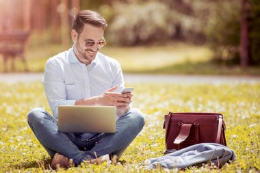 Genç adam onun laptop ile Park çimlerde oturuyor. Serbest çalışma kavramı. O telefonda mesaj yazmak.