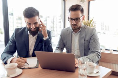Ofis salonunda masada oturan, smartphone üzerinde konuşmak ve dizüstü bilgisayar kullanarak iki işadamı