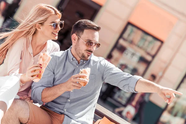 幸せな愛情のあるカップルの笑いと市のベンチでサンドイッチを食べる — ストック写真