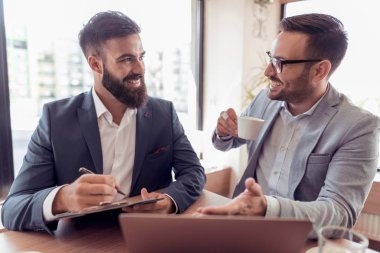 Gülümseyen, konuşurken, kahve içme, ofiste çalışırken dizüstü ve tablet kullanarak takım elbiseli iki genç yakışıklı iş adamları.