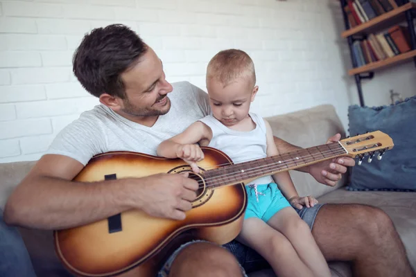Father playing on guitar and looking on his son while sitting on sofa. Father teaching his son how to play guitar.