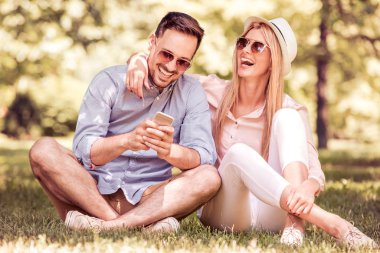 Genç çift açık bir parkta otururken akıllı telefon kullanarak. İnsanlar, aşk, eğlence ve teknoloji kavramı.