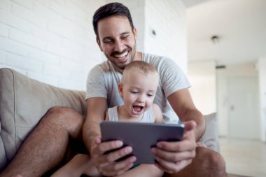 Mutlu baba ve küçük oğlu kanepede oturan ve tablet cihaza bir şey izliyor.