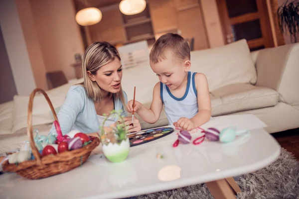 Young mother with son painting Easter eggs in living room