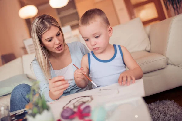 Young mother with son painting Easter eggs in living room