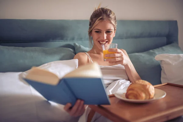 Happy young woman reading book and drinking juice in bed in morning.