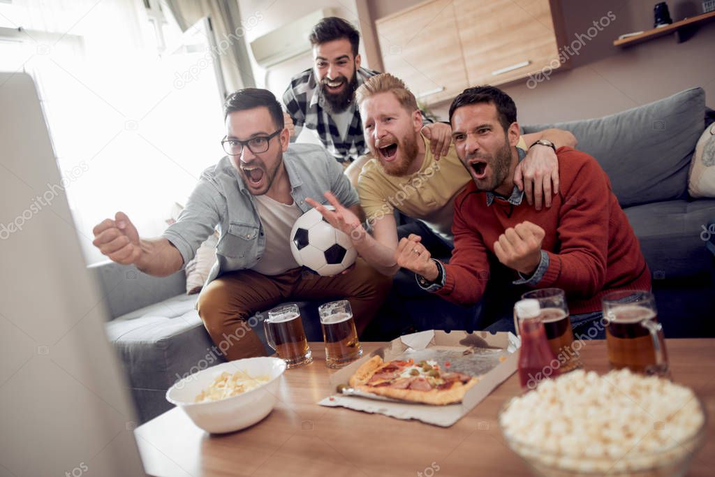 Soccer fans watching game in living room, drinking beer and eating pizza.