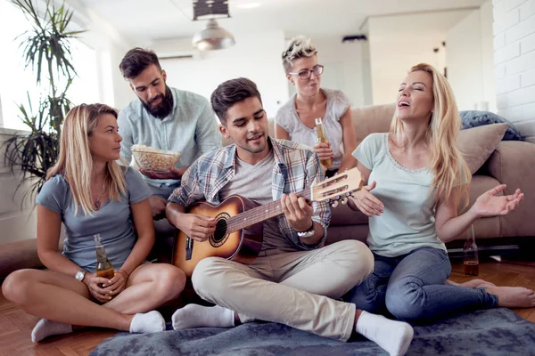 Group of friends having fun in living room in their home-Happy young people playing music with guitar,singing and laughing together .