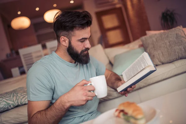 Молодой Человек Сидит Диване Читает Книгу Держа Чашку Кофе — стоковое фото