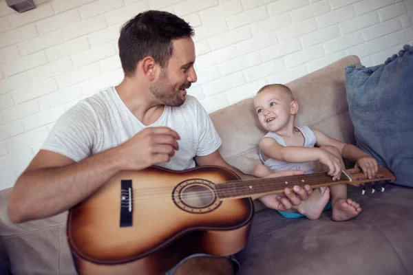 Father playing on guitar and looking on his son while sitting on sofa.Father teaching his son how to play guitar.