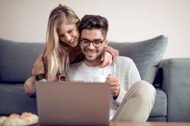 Güzel genç çift evde kanepede otururken gülümseyen bir dizüstü bilgisayar kullanarak. Çocuk bardağı götürüyor.