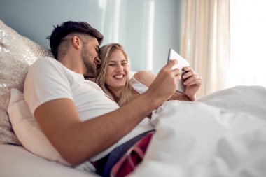 Mutlu genç çift yatakta yatıyordu ve tablet kullanma.
