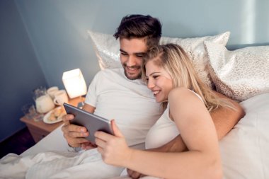 Mutlu genç çift yatakta yatıyordu ve tablet kullanma.