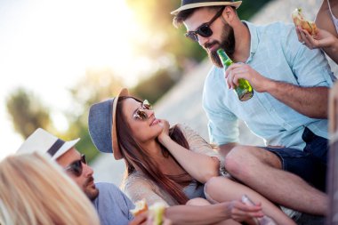 Bir grup genç, plajda piknik yaparken sandviç yiyip bira içiyor..