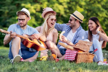 Mutlu genç arkadaşlar parkta piknik yapıyorlar. Mutlular, eğleniyorlar, gülümsüyorlar ve gitar çalıyorlar.