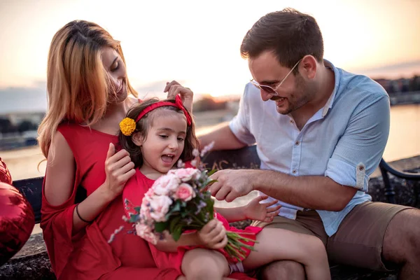 川のほとりのベンチに座っている若い家族 花の花束を持っている女の子 — ストック写真