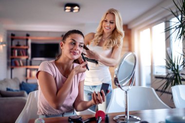 Masada makyaj yapan gülümseyen genç kız ve saçını tarayan kız arkadaş.