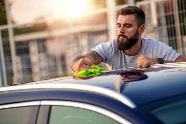 Genç adam dışarıda arabasını temizliyor. Mikrofiberli bir adam arabanın cilasını siliyor..