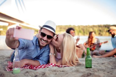 Mutlu genç çift pikniğin tadını çıkarıyor ve sahilde selfie çekiyor..