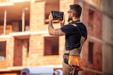 İş, inşaat, teknoloji ve insan konsepti - inşaat alanında duran mühendisin arka planı ve tabletle fotoğraf çekmek.