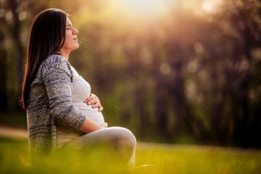 Hamile bir kadın oturmuş karnını tutuyor ve doğanın tadını çıkarıyor..