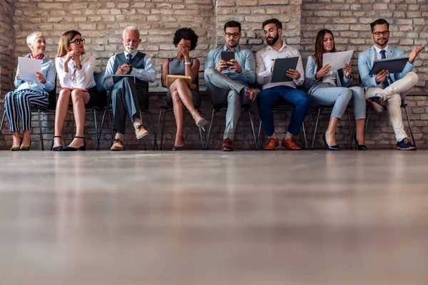 Line of  people different ages sitting by wall while waiting their turn for job interview.