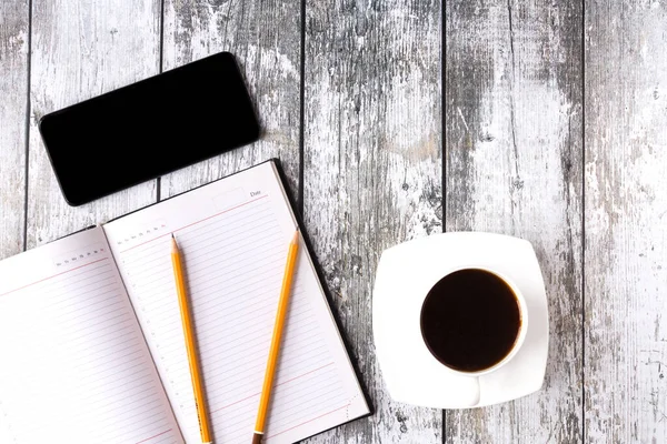 Smartphone with black screen on a rough wooden background with cup of black coffee. An opened notebook with two pencils on it lie next to them. Business meeting or coffee break in the office