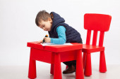 Aranyos kisfiú rajz, piros asztal és szék, fehér háttér. Gyermekkori oktatási koncepció