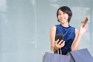 Genç ve güzel Asyalı bir kadın cep telefonundan kredi kartıyla alışveriş yaparken kollarında renkli alışveriş torbalarıyla gülümsüyor..