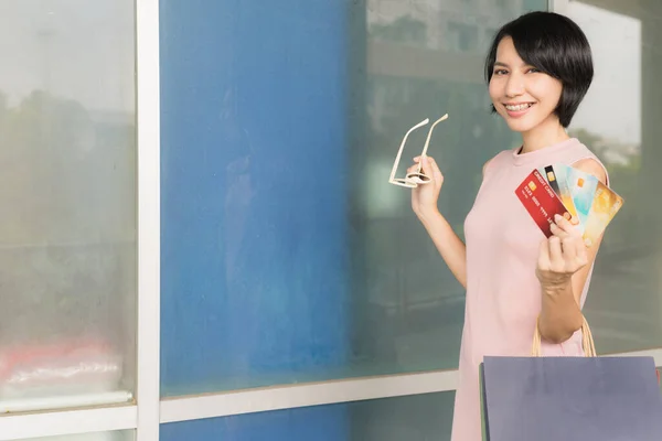 Beautiful asian shopping girl wearing pink dress, carrying a shopping bag and showing credit cards