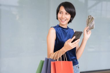 Genç ve güzel Asyalı bir kadın cep telefonundan parayla alışveriş yaparken kollarında da gülümseyen yüzüyle alışveriş torbaları tutuyordu..