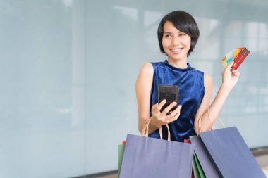 Genç ve güzel Asyalı bir kadın cep telefonundan kredi kartıyla alışveriş yaparken kollarında renkli alışveriş torbalarıyla gülümsüyor..