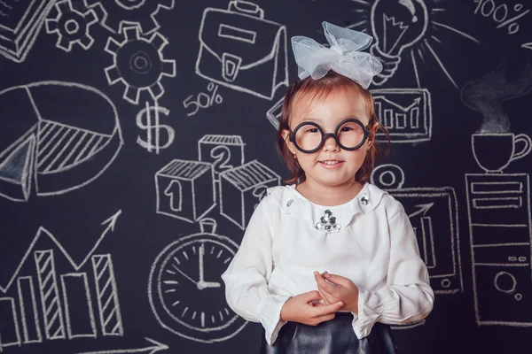 Маленька розумна дівчинка в окулярах на фоні стіни з діловим або шкільним зображенням — стокове фото