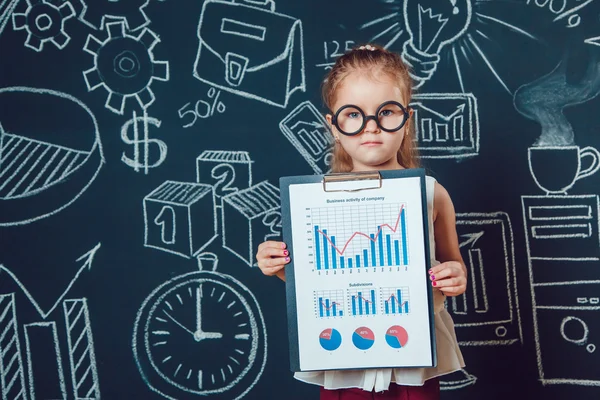 The little smart girl in glasses shows graphics companys business activity on a background of wall with picture — Stock Photo, Image