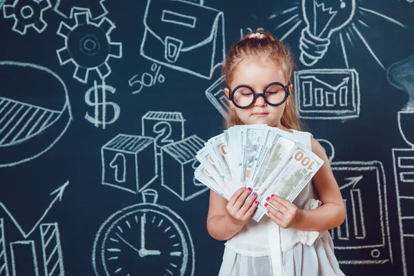 La niña inteligente en gafas con dinero en un fondo de la pared con la imagen — Foto de Stock